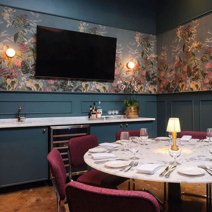 Private dining room with a round marble table set for eight, plum chairs, teal wainscoting and floral wallpaper, small table lamp, built-in bar with sink and wine fridge, and a wall-mounted TV