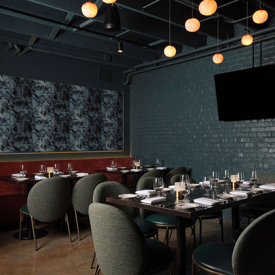 Moody BKS dining room with dark teal brick walls, red banquette, globe pendant lights, sage upholstered chairs around set dark-wood tables, and a wall-mounted TV.