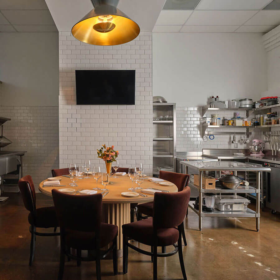 Round table set with stemware and burgundy chairs inside a working kitchen—white subway tile, stainless prep stations, open shelves, TV, and a gold-lined pendant—chef’s-table seating