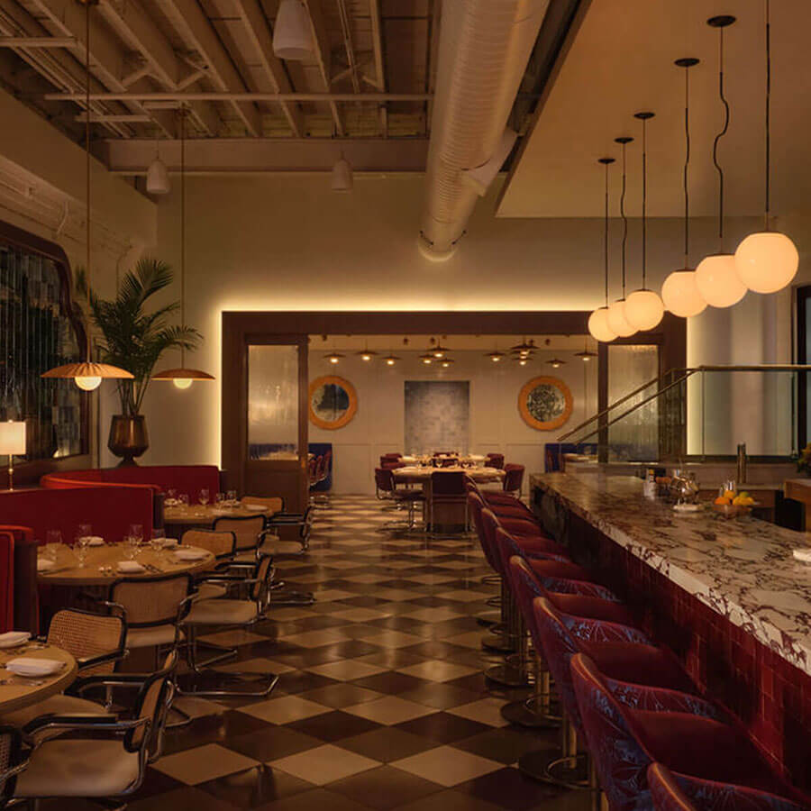 Low-lit space where a marble bar with globe pendants faces deep red banquettes and rattan bistro chairs on a checkerboard floor, opening to round tables in the adjoining room.